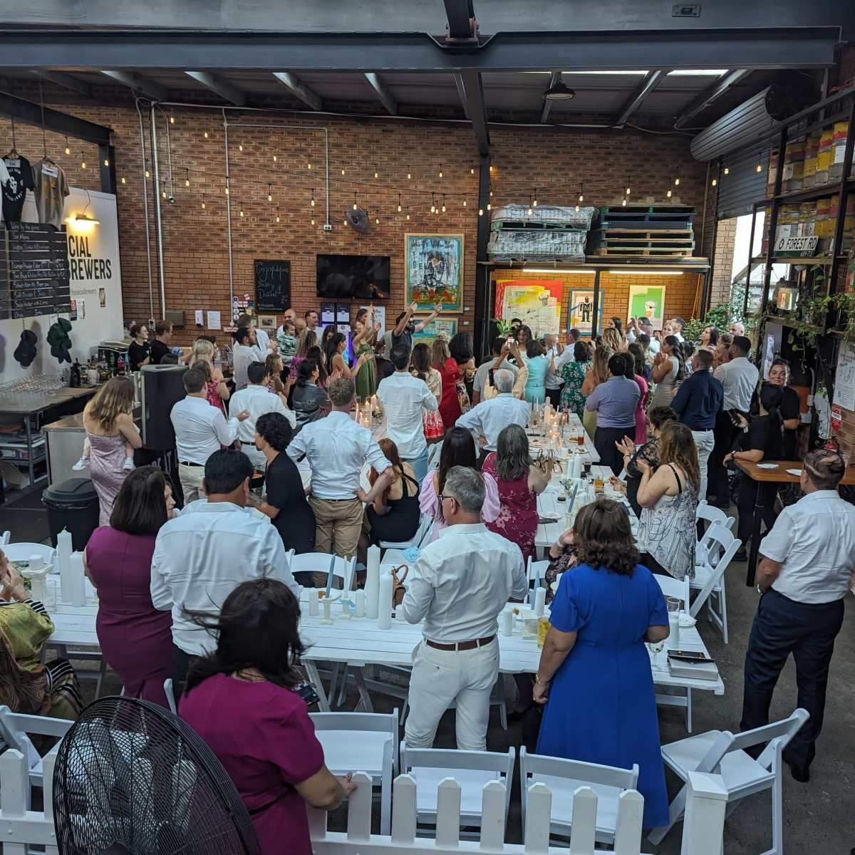 A large gathering of people in formal attire is taking place in a spacious venue with exposed brick walls and a bar area.