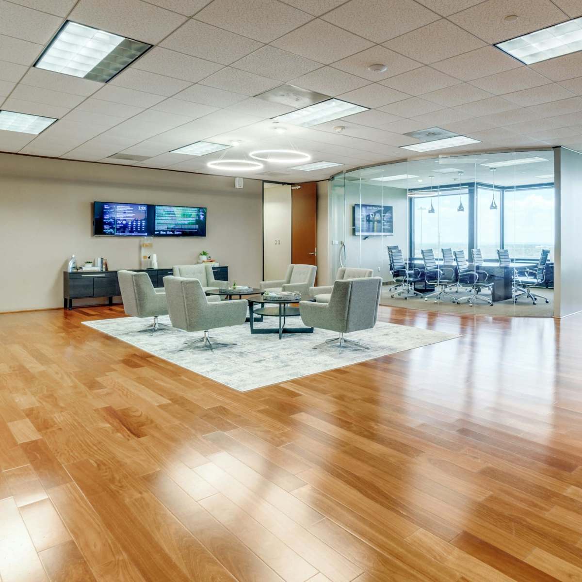A modern office space featuring a seating area with gray chairs, a conference room in the background, and large windows with a view.