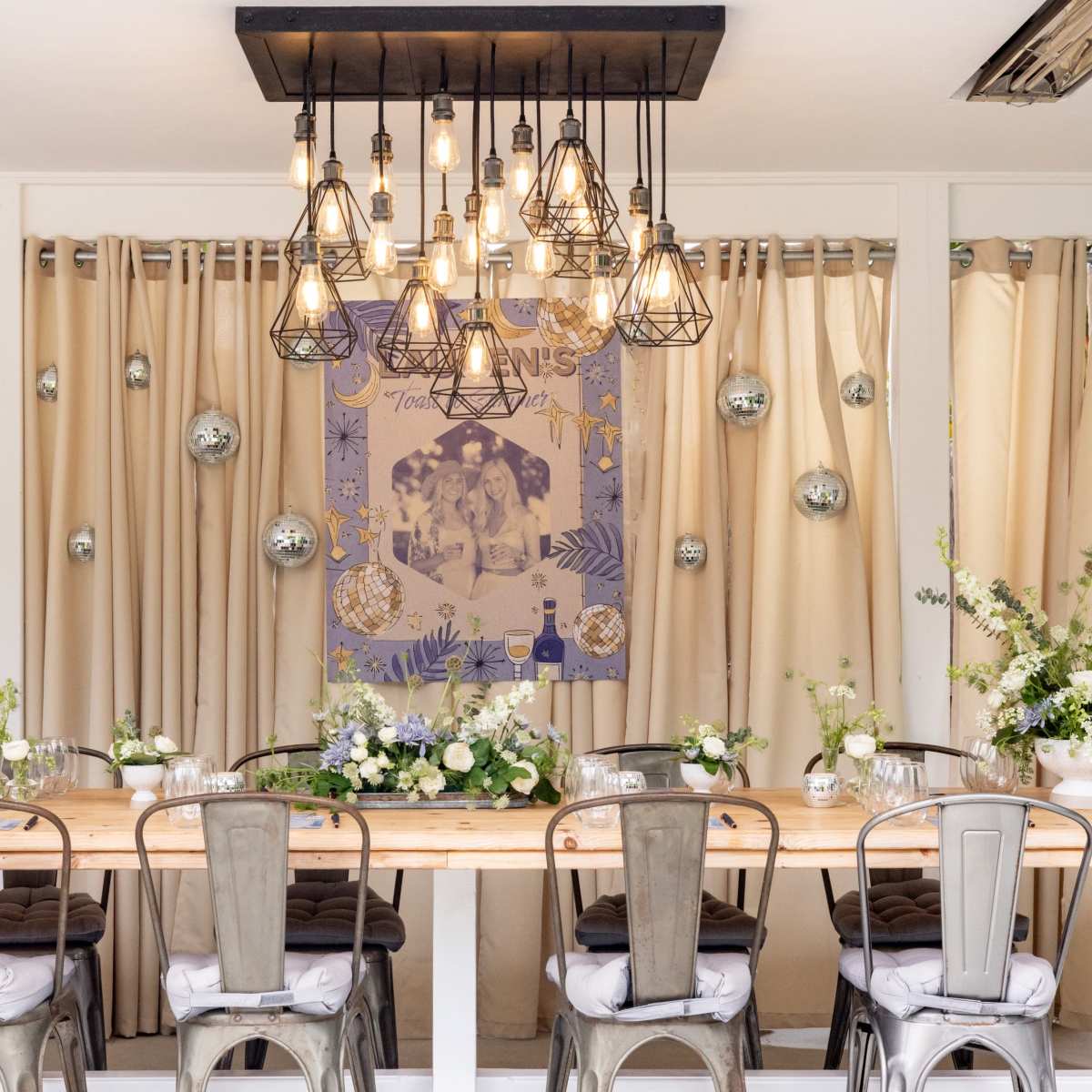 A long wooden dining table with metal chairs is set beneath a modern chandelier, surrounded by white drapes and adorned with floral arrangements.