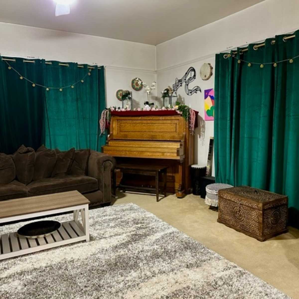A cozy living room with a brown sofa, a wooden piano, green curtains, and decorative items on a table.