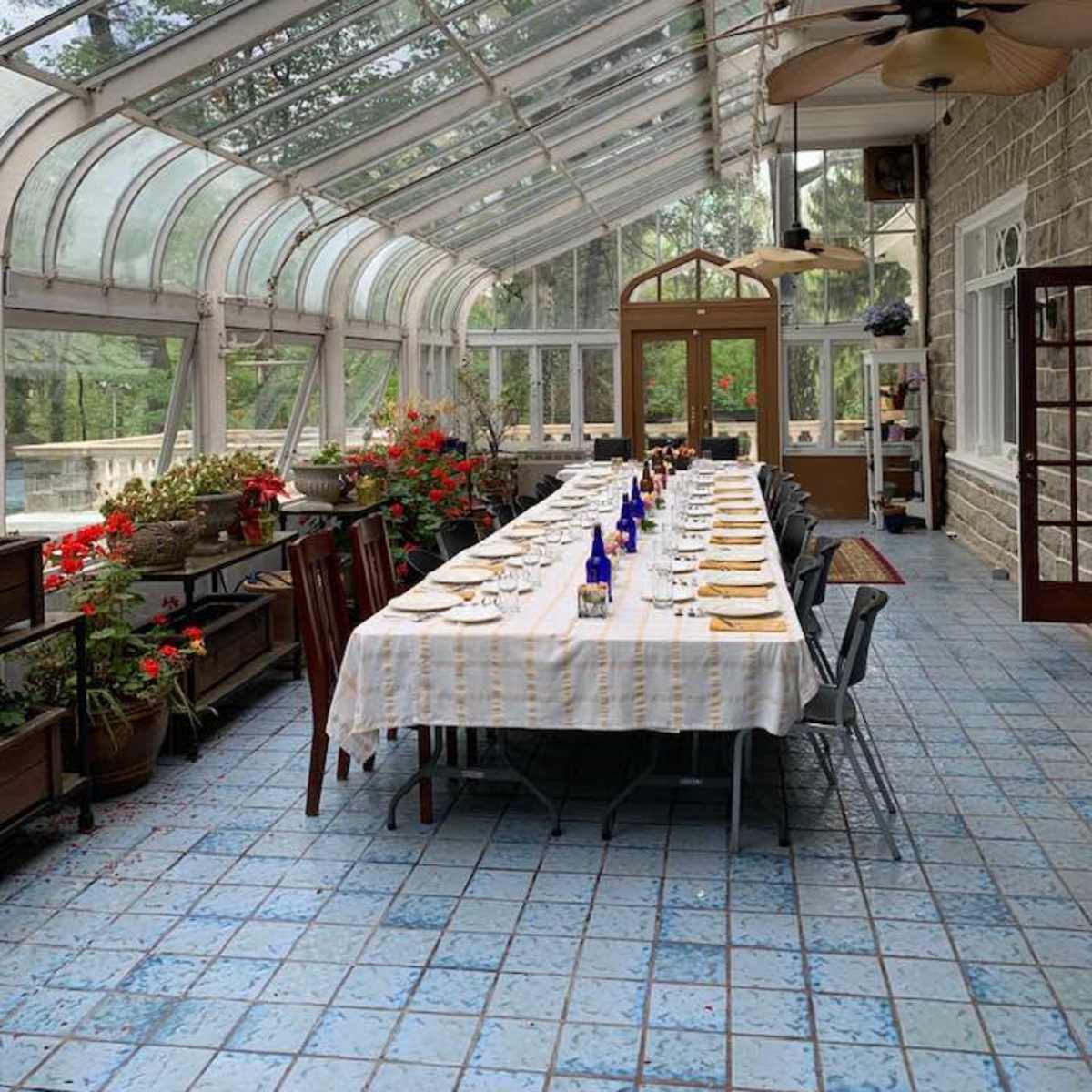 A long dining table is set under a glass-roofed structure with potted plants and flowers along the sides.