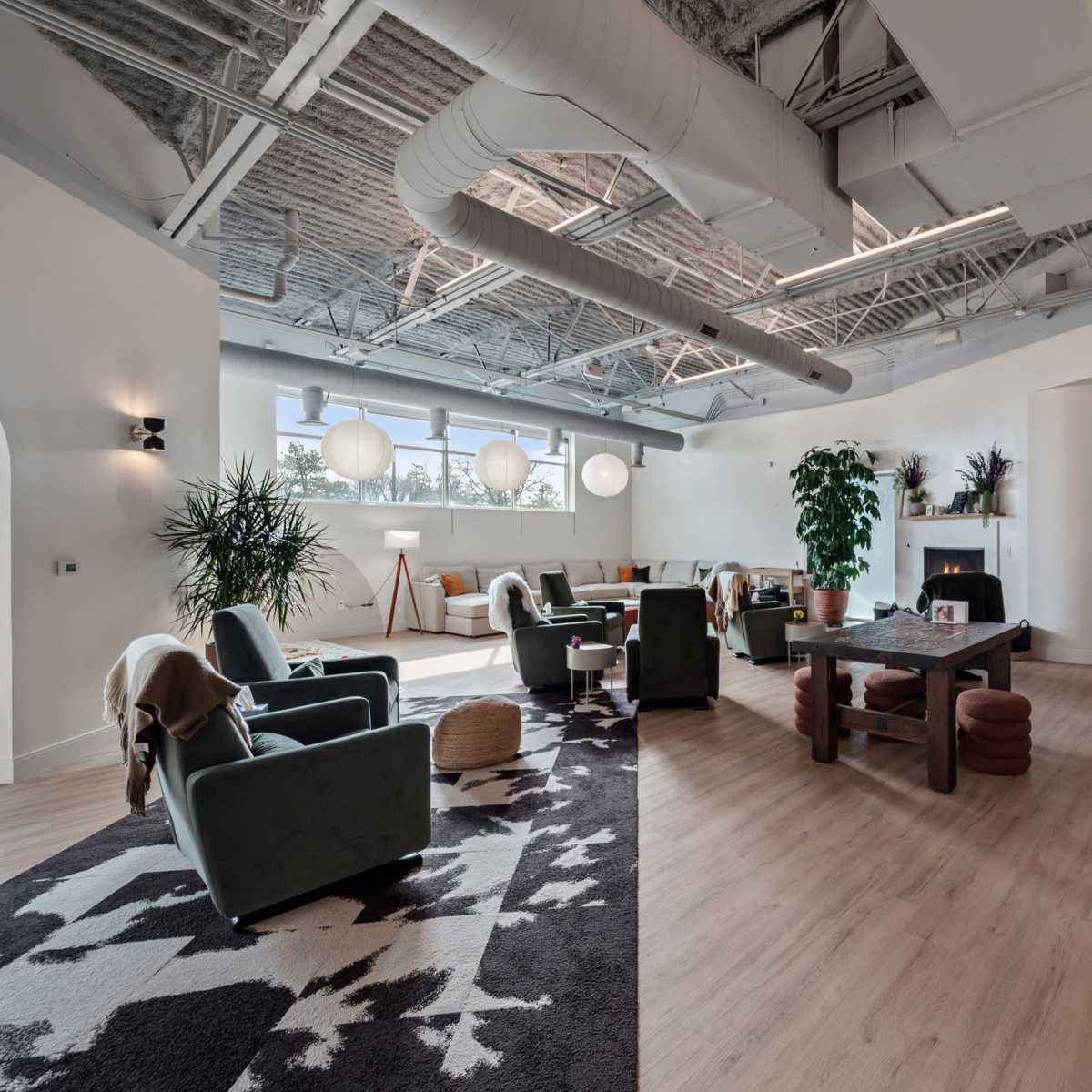 A modern lounge area featuring comfortable seating, large windows, plants, and a communal table.