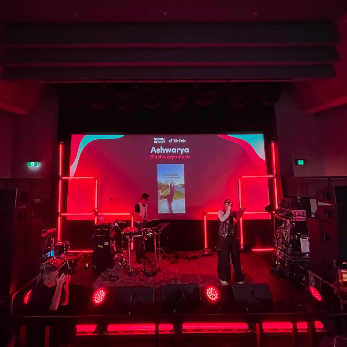 A performer stands on stage in front of a large screen displaying their name and social media handle, with instruments set up for a band in the background and red lighting illuminating the scene.