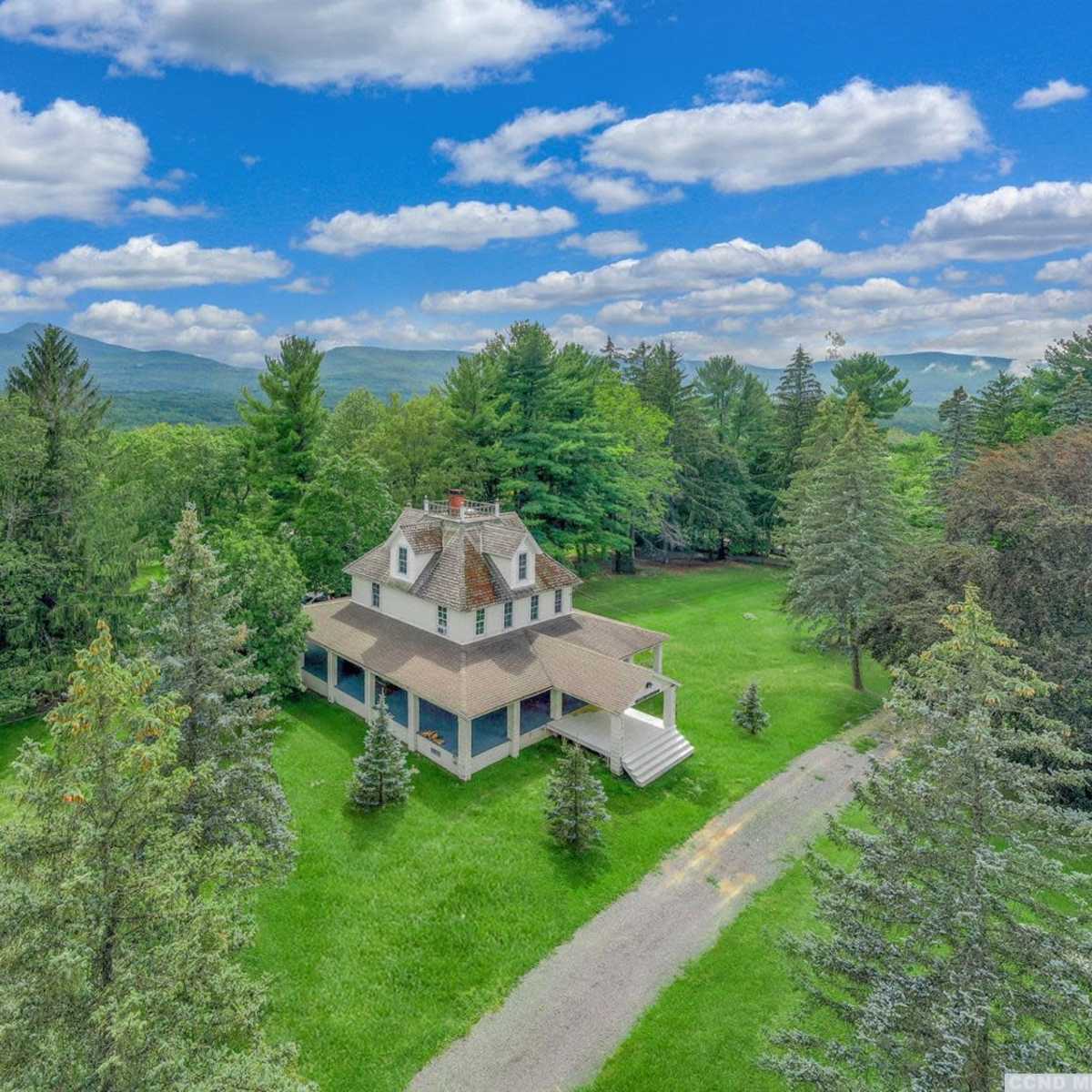 A large, two-story house surrounded by lush green trees and a gravel driveway, set against a backdrop of mountains and a partly cloudy sky.