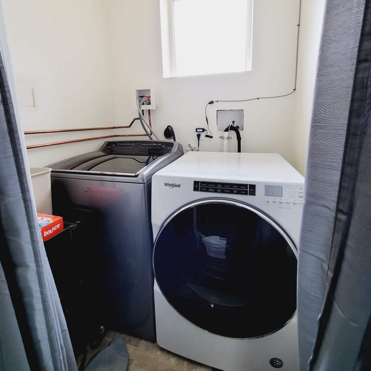 A laundry room with a washing machine and a dryer next to each other, a small window providing natural light, and utility connections on the wall.