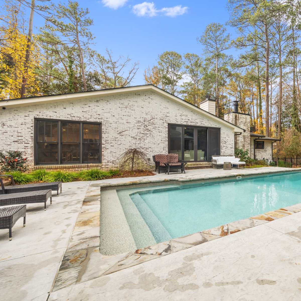 A backyard with a swimming pool surrounded by a stone patio and vegetation, adjacent to a house with large windows.
