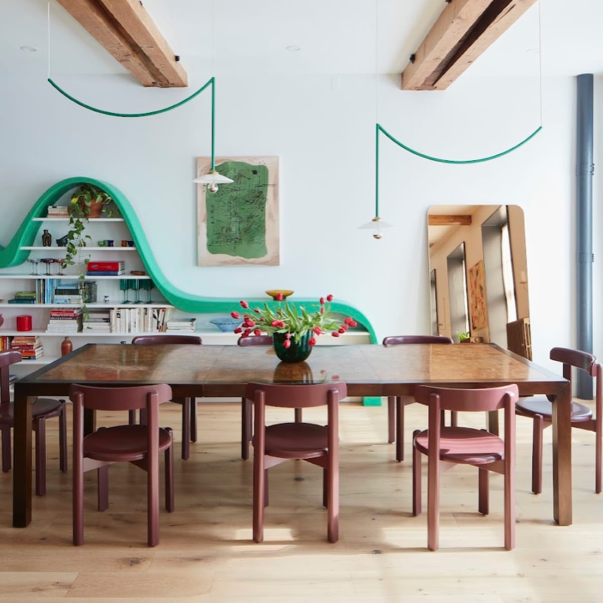 A modern dining room features a long wooden table surrounded by chairs, with green decor elements and open shelving in the background.