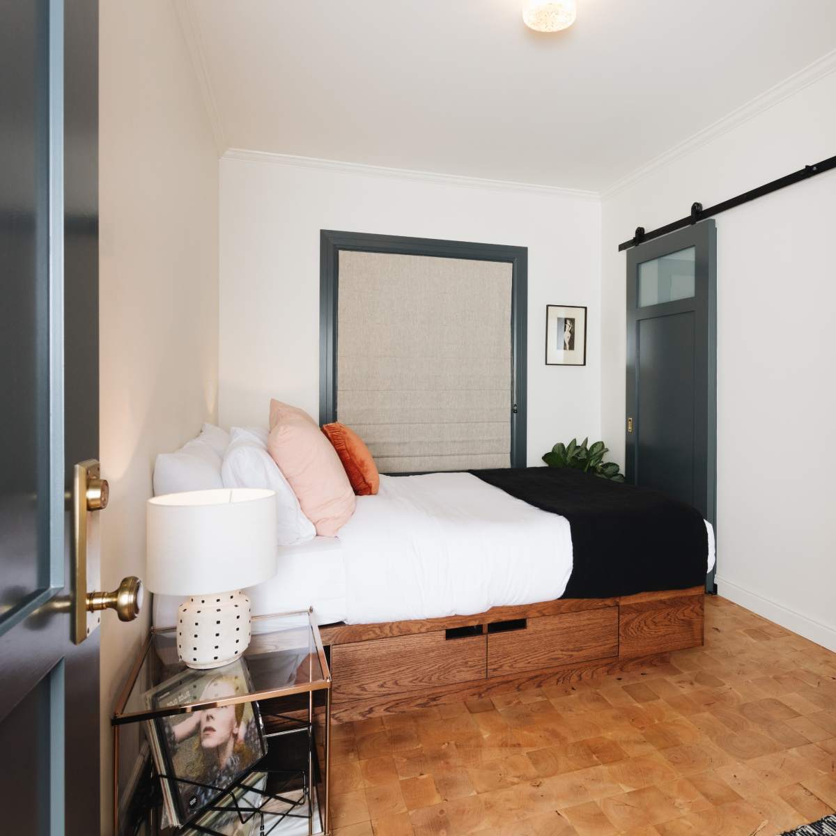 A modern bedroom features a bed with white linens and orange accents, a wooden storage base, and sliding barn door, along with a glass shower visible in the background.