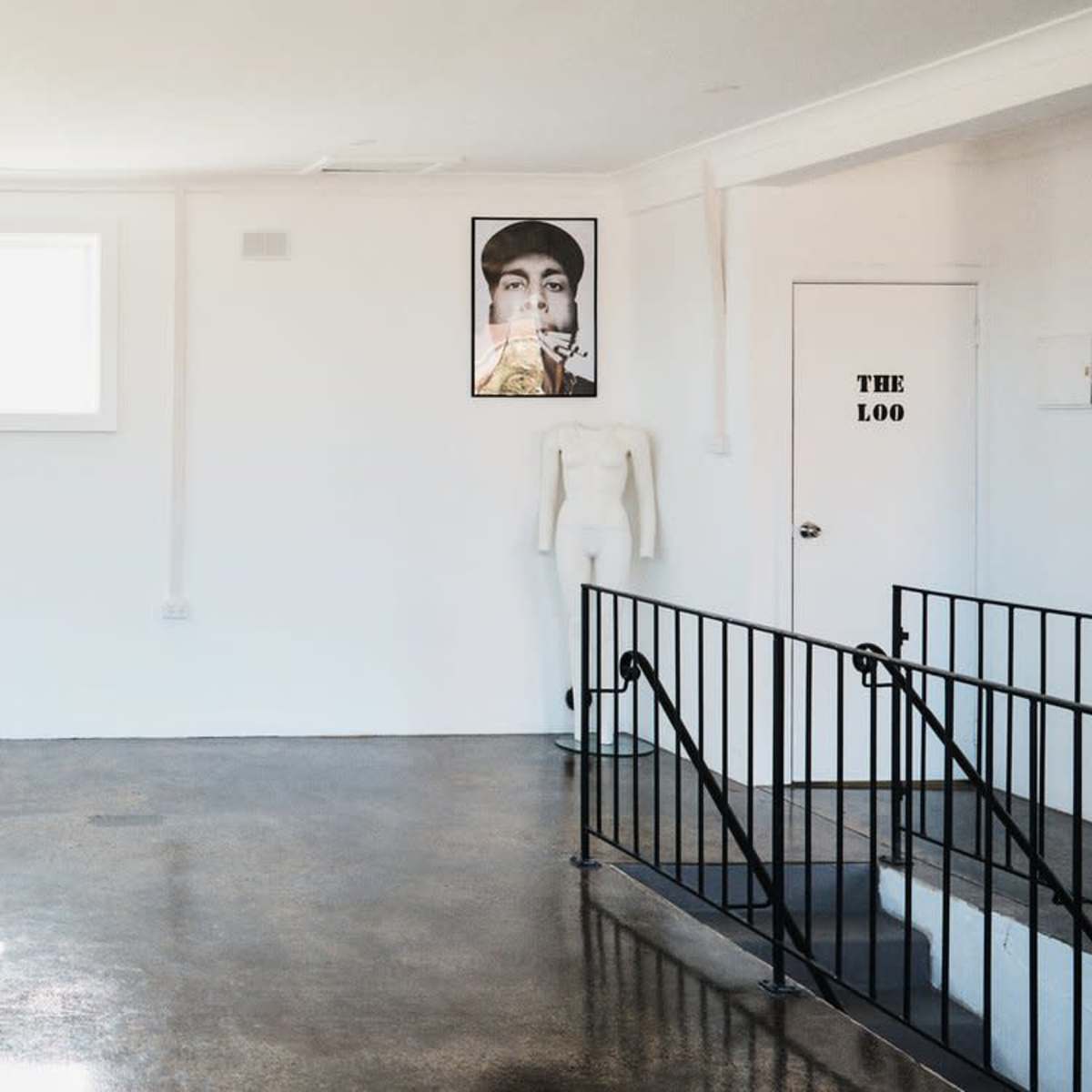 A spacious, empty room with a black table, a window, and a staircase leading to a lower level, featuring a mannequin and a framed photo on the wall.