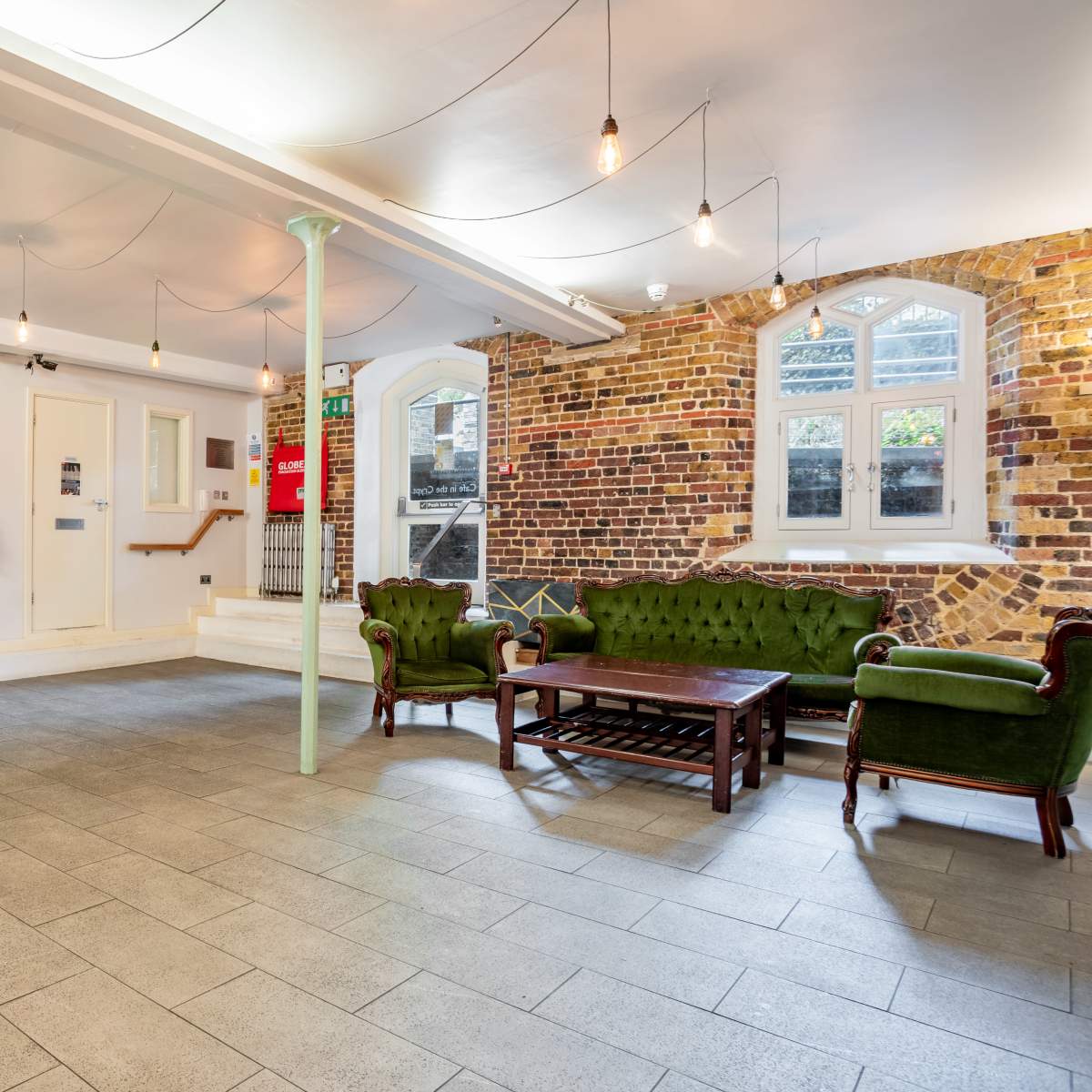 A spacious interior with stone flooring, a green velvet sofa set, and exposed brick walls, illuminated by hanging light bulbs.