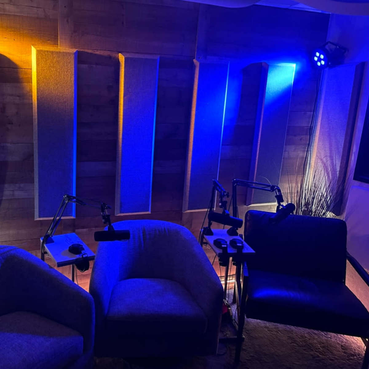 A cozy recording studio with two blue armchairs and soundproof wall panels illuminated by warm yellow and cool blue lights.