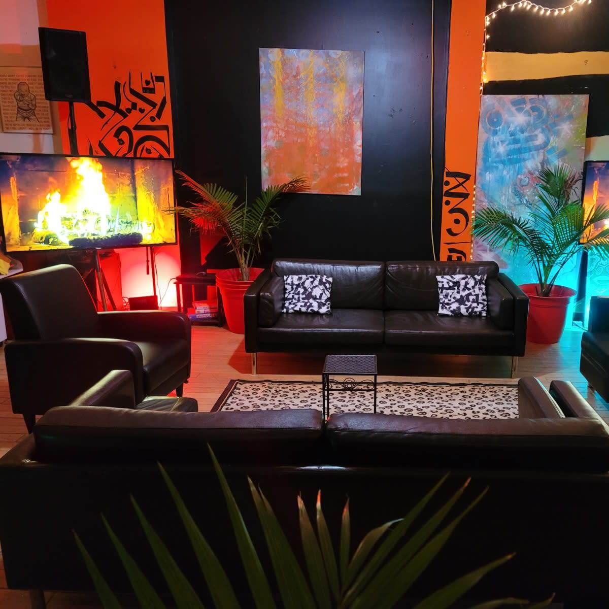 The scene features a modern lounge area with black leather furniture, two large televisions displaying flames, and colorful backgrounds illuminated by lights, complemented by potted plants.
