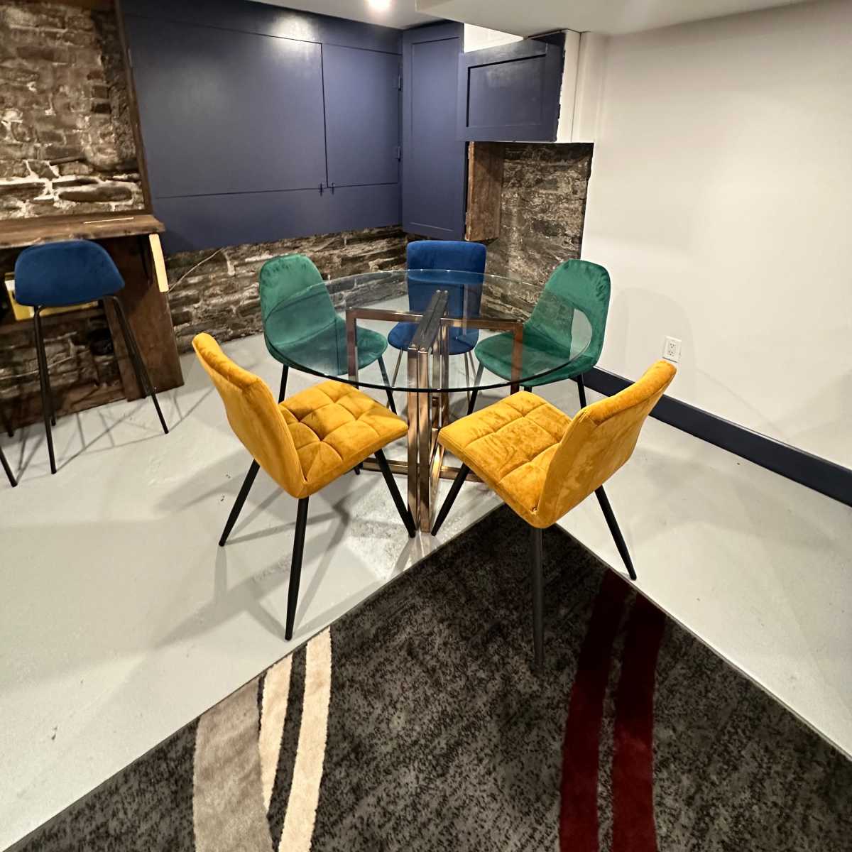 A modern seating area features a glass table surrounded by four colorful chairs on a patterned rug.