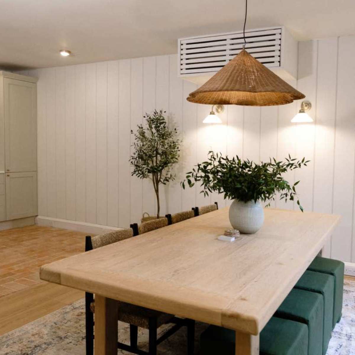 A spacious dining area features a wooden table surrounded by chairs, with a potted plant centerpiece and a textured rug on the floor.