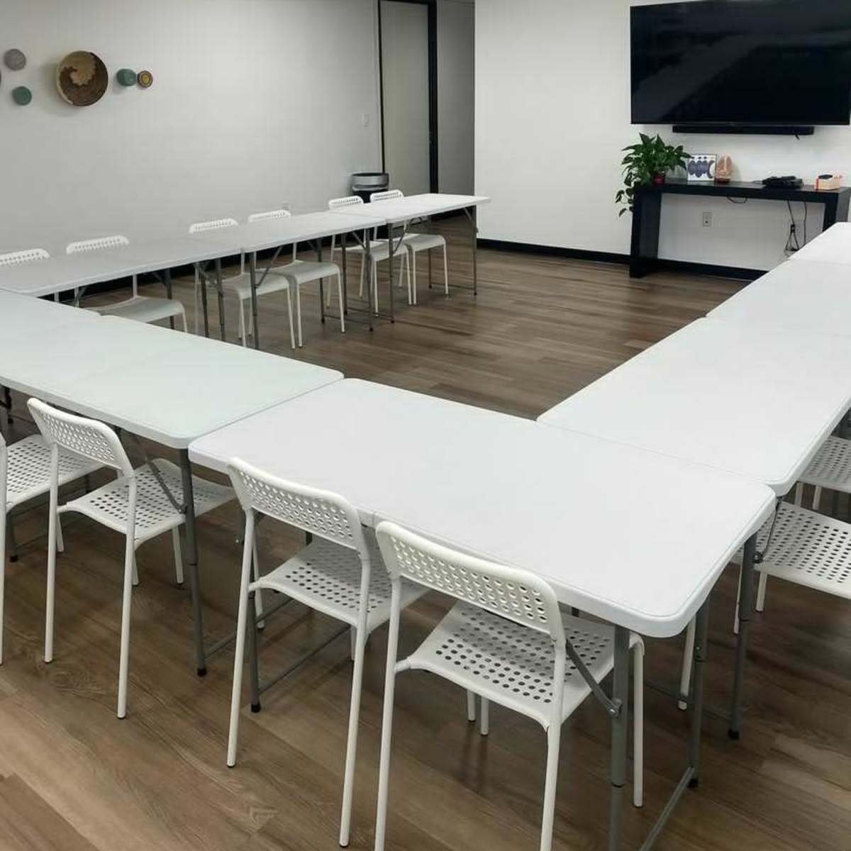 A conference room features a U-shaped arrangement of white tables surrounded by chairs, with a large screen mounted on the wall.
