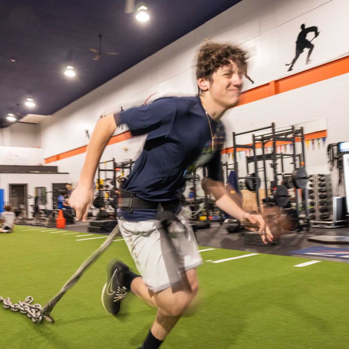 A young man runs on turf while attached to a training harness with chains in a gym equipped with weightlifting stations and fitness equipment.