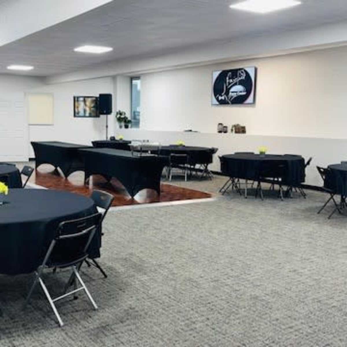 A spacious event room features several round tables covered with black tablecloths, arranged with small yellow flower centerpieces.