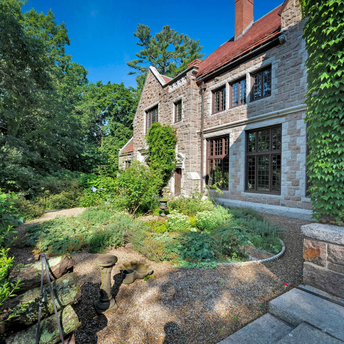 A stone house with a red roof is surrounded by lush greenery and a garden pathway.