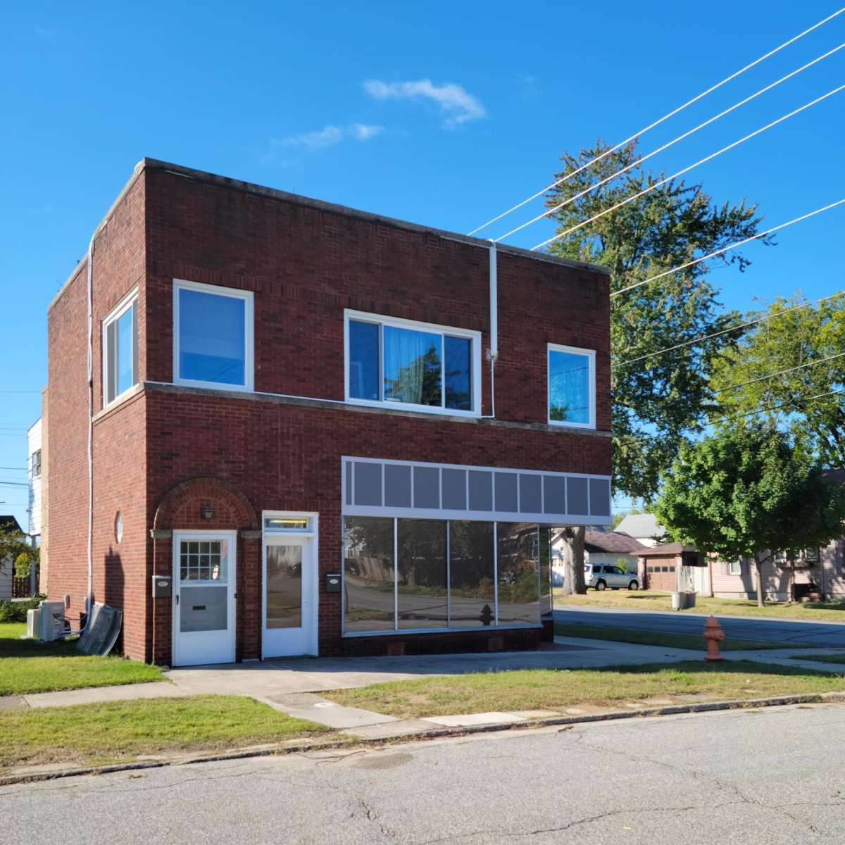 A two-story brick building with large windows and a glass storefront sits on a corner lot, facing a residential street.