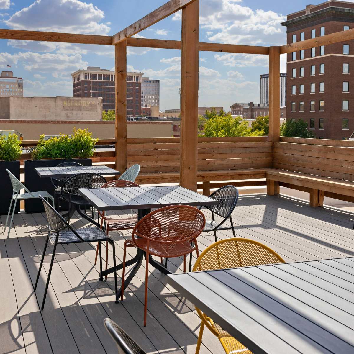 A rooftop terrace with wooden beams, metal chairs, and tables, overlooking a city skyline.