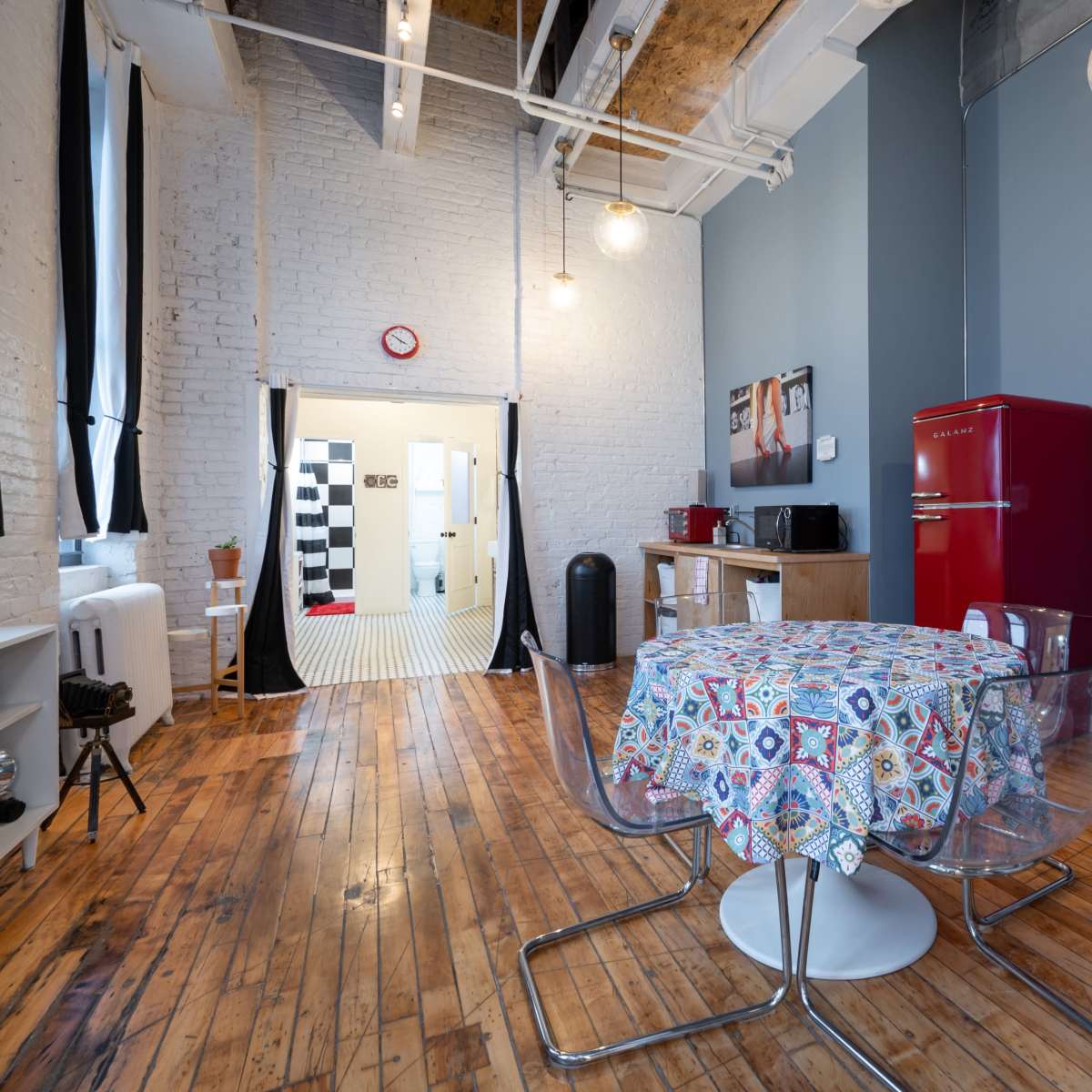 A modern kitchen area with a round table, transparent chairs, and a red refrigerator, featuring a doorway that leads to a black-and-white tiled bathroom.