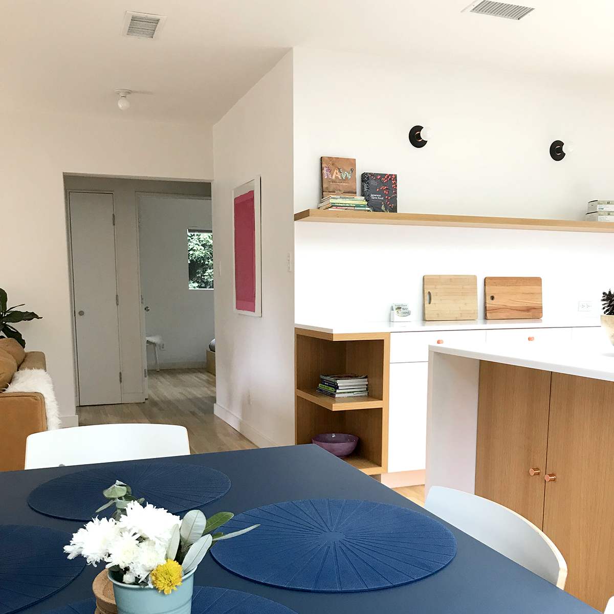 A modern dining area features a table with blue placemats, surrounded by white chairs, and a view into a well-lit kitchen and adjoining room with neutral decor.