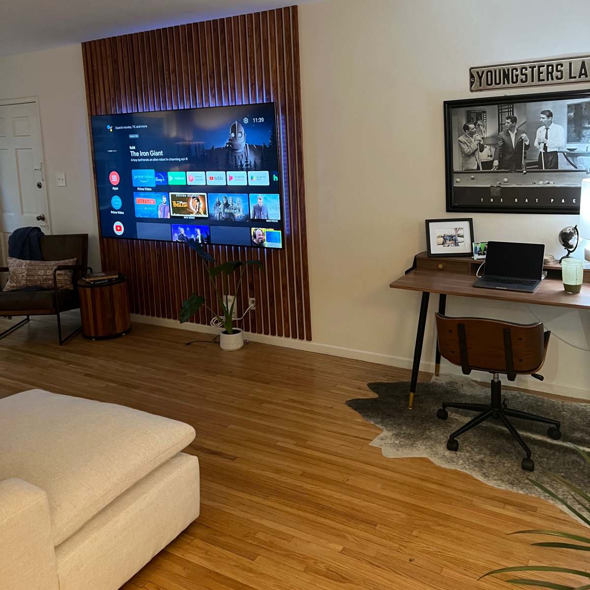 A modern living room features a wall-mounted television and a desk area with a chair, surrounded by wooden accents and a rug on the floor.