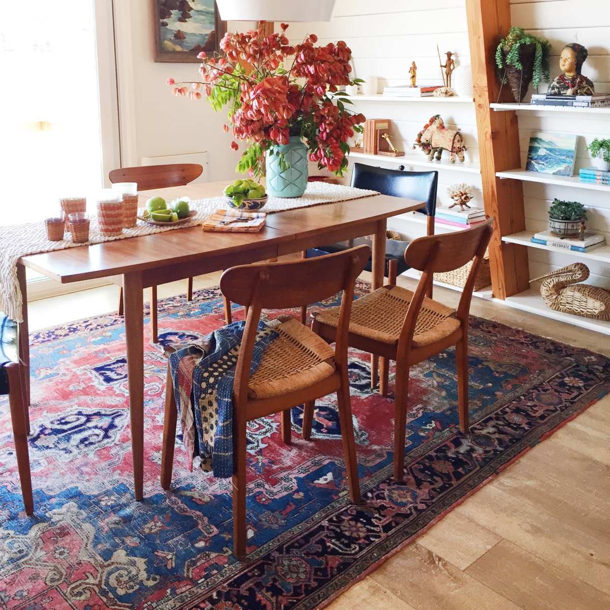 A dining area features a wooden table with four chairs, a large vase of flowers as the centerpiece, and shelves displaying various decorative items.