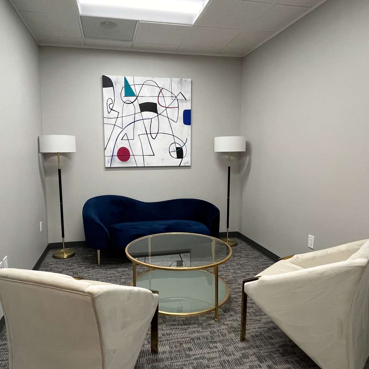 A small meeting room features a blue couch, two white chairs, a glass coffee table, and a colorful abstract painting on the wall.
