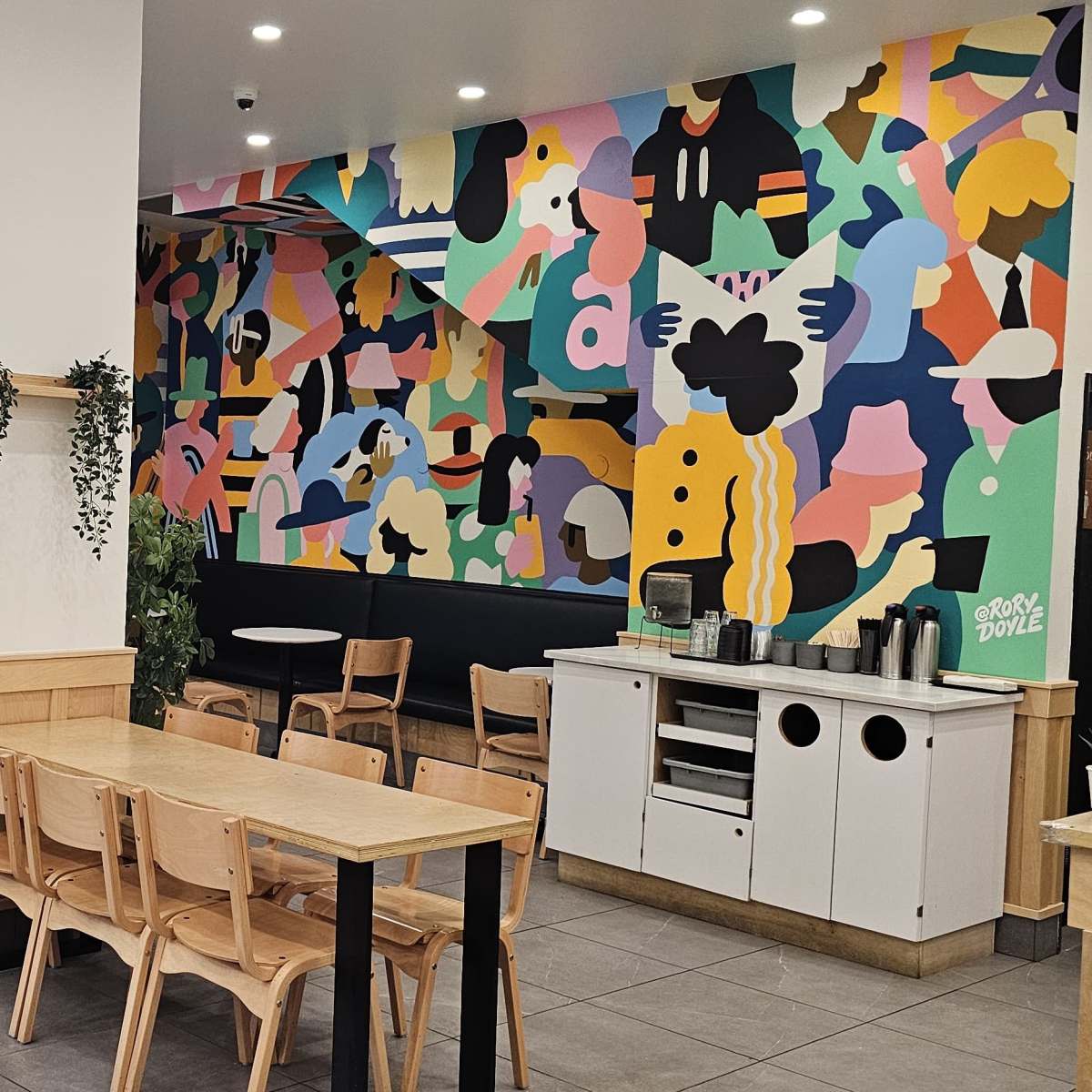 A brightly decorated café features a vibrant mural on one wall, wooden furniture, and a display of pastries in the foreground.