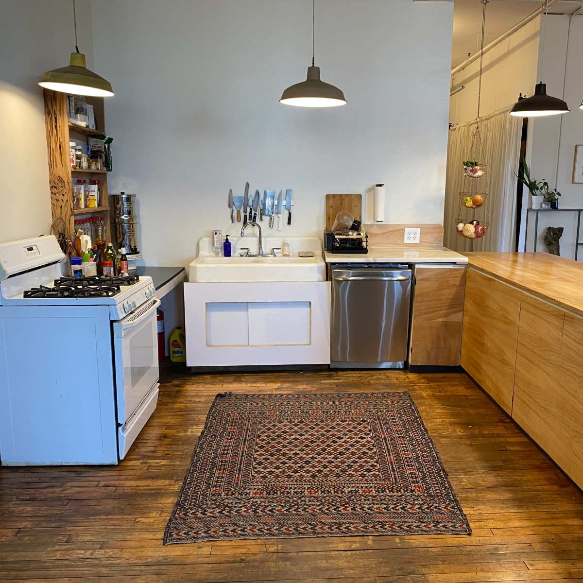 A modern kitchen features a white sink, a gas stove, a stainless steel dishwasher, wooden cabinetry, and a patterned rug on the floor.