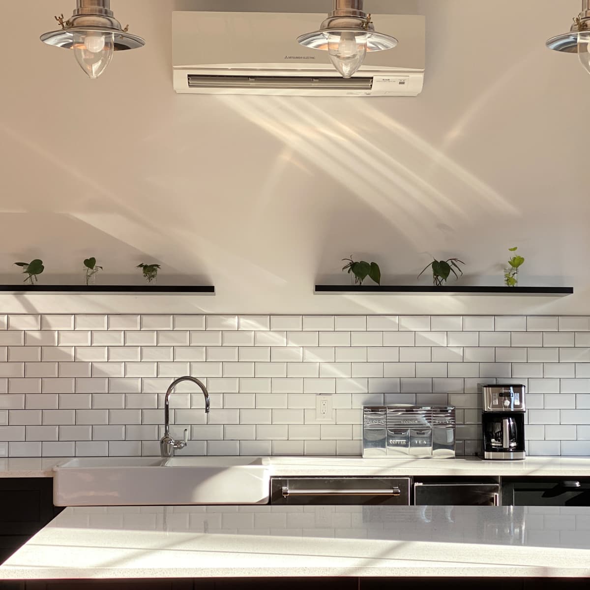 A modern kitchen with white tiles, a large sink, a stovetop, and shelves displaying small plants under overhead lighting.