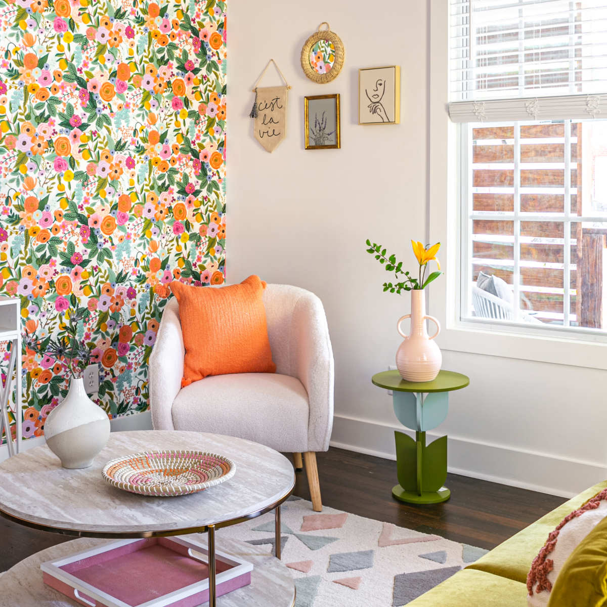 A bright living room features a floral accent wall, a white armchair with an orange cushion, a round coffee table, and a green side table with a vase and a yellow flower.