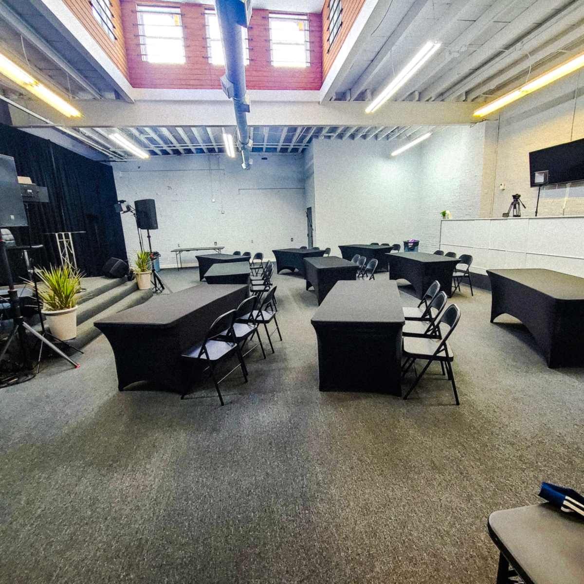 A spacious room set up for an event, featuring several tables with black covers and chairs arranged throughout, along with audio equipment and plants.