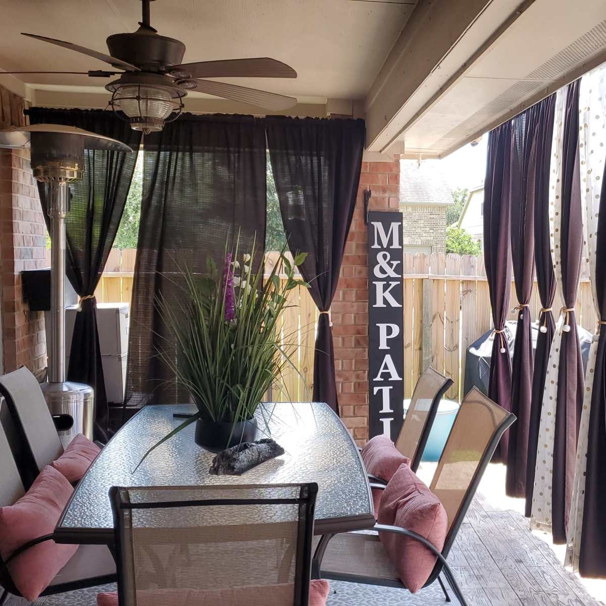 A covered patio area with a glass table, chairs, a ceiling fan, and dark curtains framing an adjacent space.