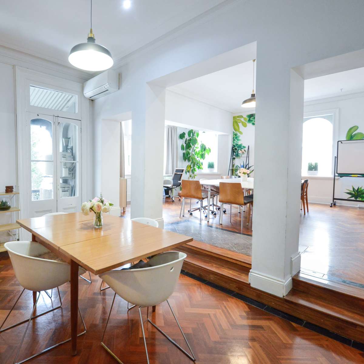 A bright, open dining area featuring a wooden table with white chairs, leading into a well-lit office space with plants and modern furniture.