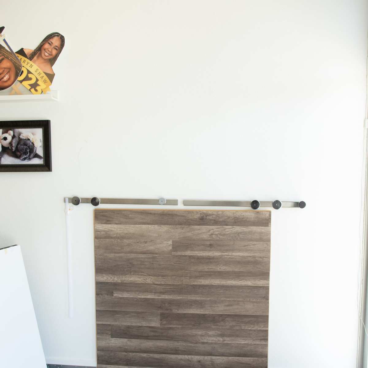 A wall with a wooden panel mounted on a track, alongside a few framed pictures and a cardboard cutout on a shelf.
