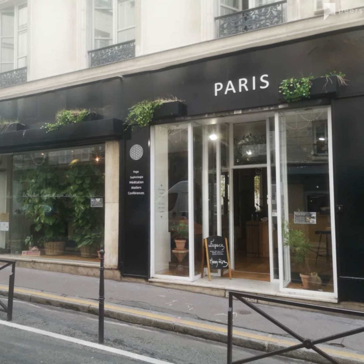 A café with large glass windows and outdoor plants is located on a street in Paris.
