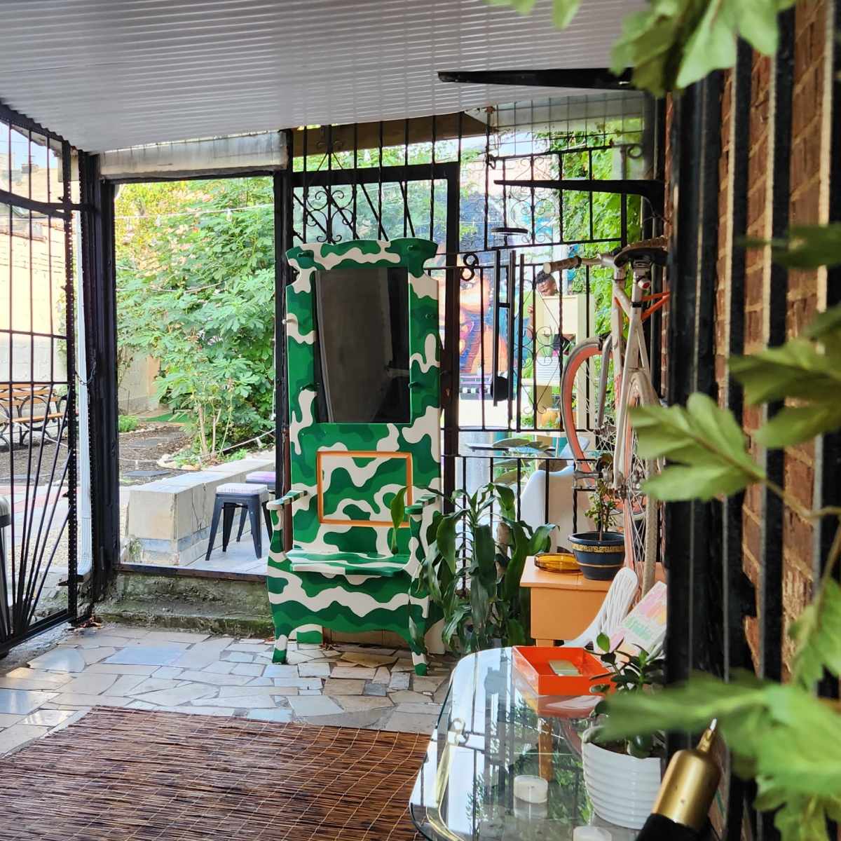 A narrow outdoor corridor with metal gates leading into a garden area, surrounded by plants and various pieces of outdoor furniture.