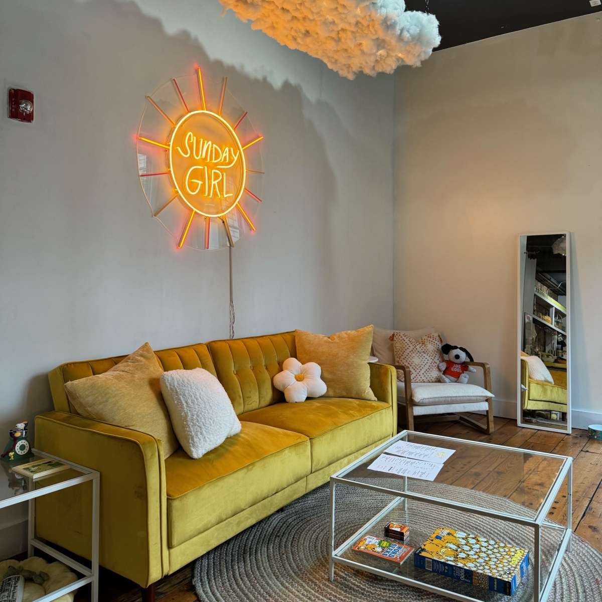 A bright yellow sofa is positioned under a neon sign that reads "SUNDAY GIRL," with a cloud-shaped decoration above it in a stylish, minimalist room.