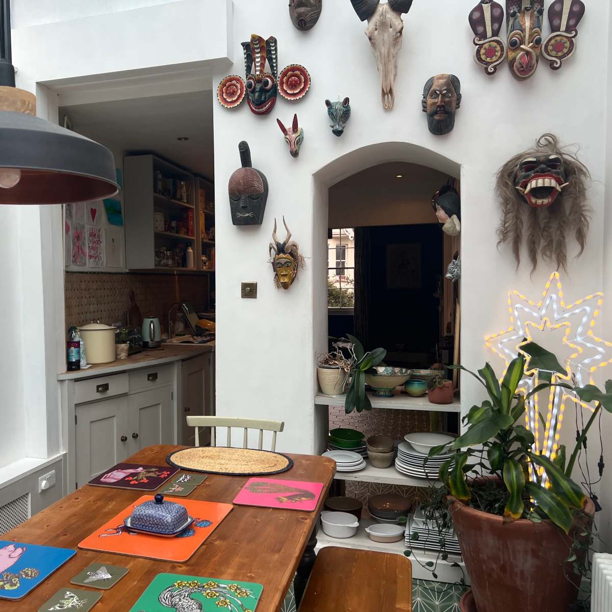 A wooden dining table is set in a brightly lit room adorned with various masks and artwork, with a kitchen visible in the background.