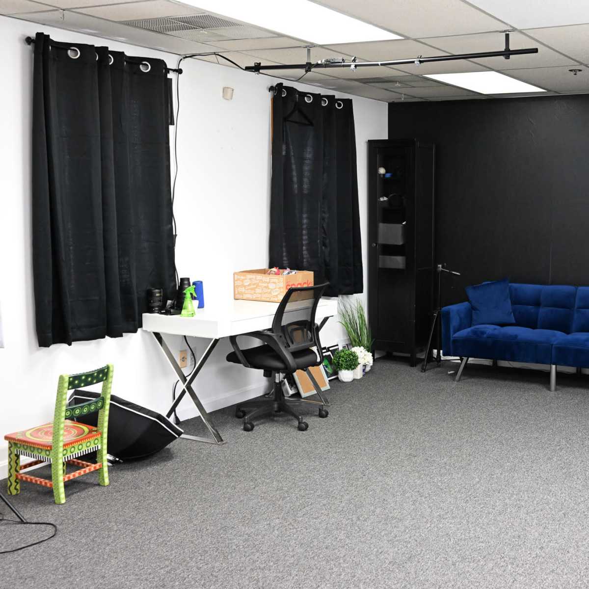 A sparse room with gray carpeting, a blue couch, a desk with a chair, two black curtains, and a few decorative plants.