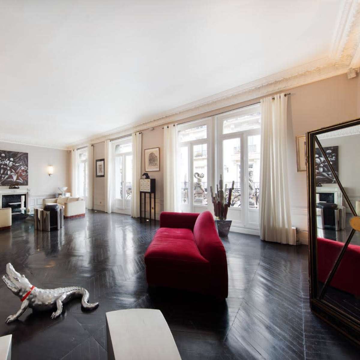 A spacious living room featuring large windows, a red couch, a metallic sculpture, and a floor with dark wood finish.