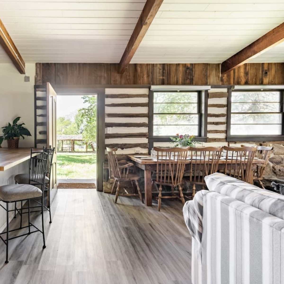 The interior of a modern cabin features a kitchen area with a bar counter, a dining table set for several people, and large windows overlooking a view of nature outside.
