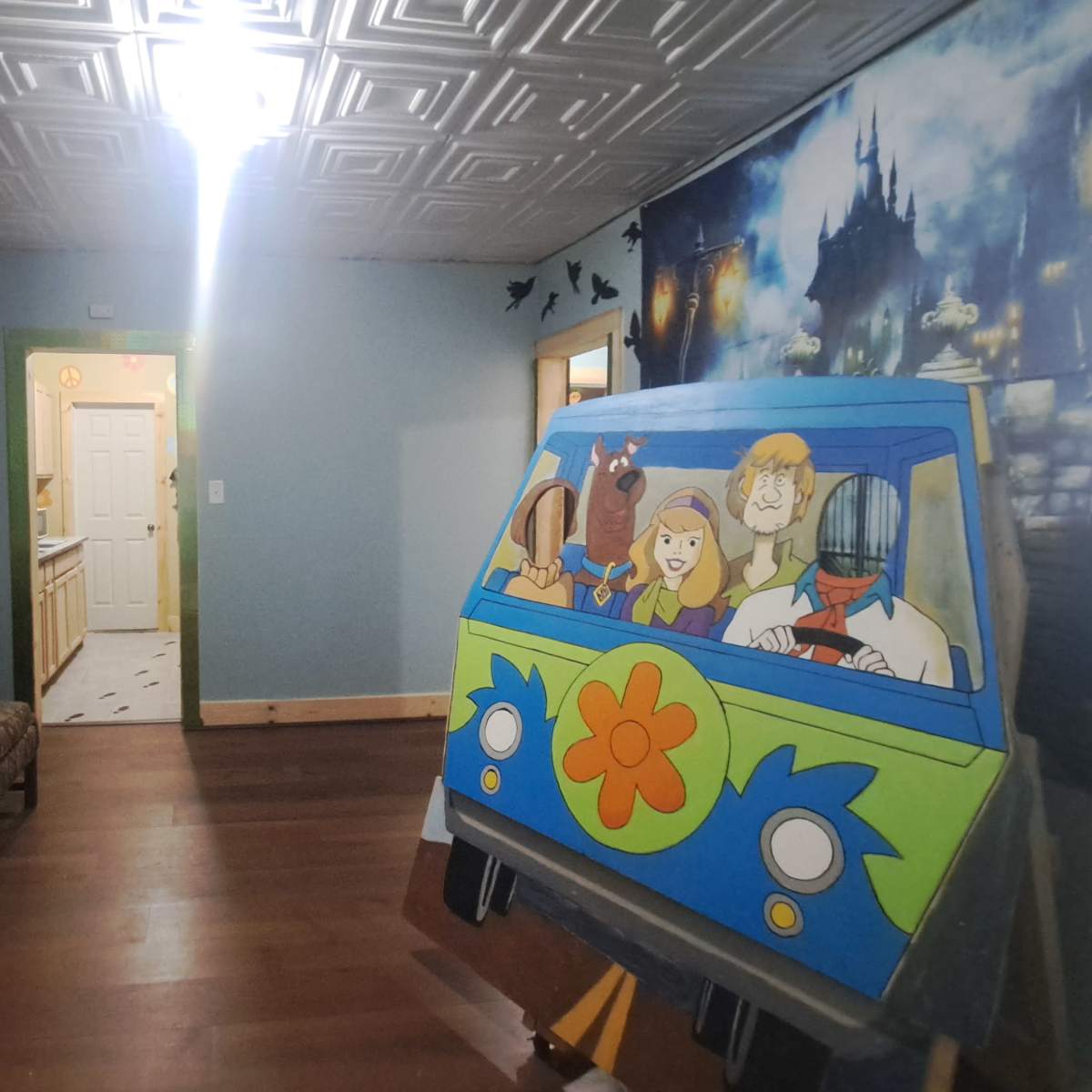 The scene features a colorful room with a cartoon-themed curtain, a patterned sofa, and a mural of a cartoon vehicle accompanied by characters on one wall, with a doorway leading to a kitchen visible in the background.