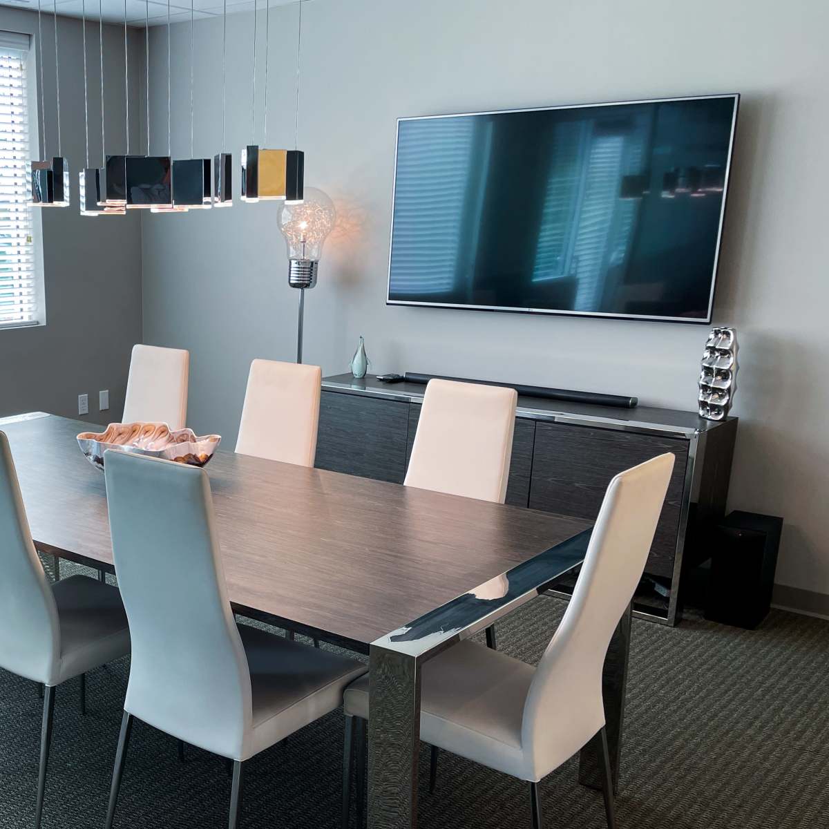 A modern dining room features a long table surrounded by eight light-colored chairs, with a television mounted on the wall and a window providing natural light.
