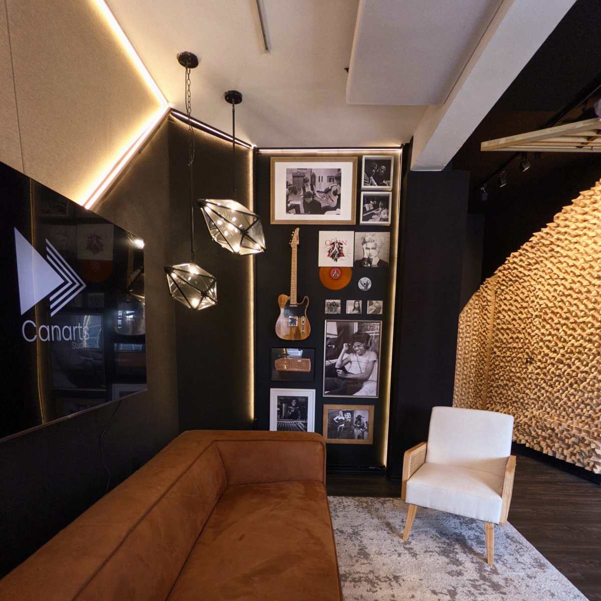 A modern interior space with a brown velvet couch, a light-colored armchair, framed black and white photographs, a guitar, and contemporary lighting fixtures against a dark wall.