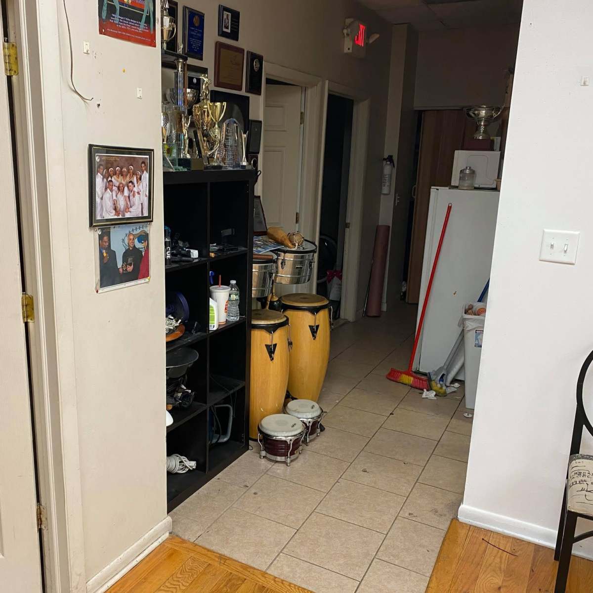 A hallway leading to a kitchen area, with shelves displaying trophies and instruments, and tiled flooring.