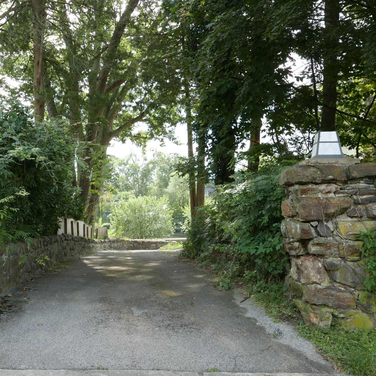 A gravel driveway lined by stone pillars leads into a wooded area flanked by trees.