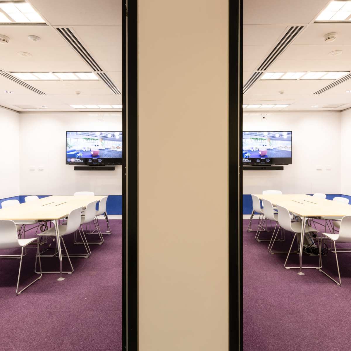 A modern conference room features a long table surrounded by white chairs, with two screens displaying content on opposite walls.
